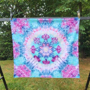 May include: A square, tie-dye textile in shades of blue, pink, and white, hanging outdoors. The pattern is symmetrical, with a central floral design. The fabric is displayed on a black metal frame, with green grass and trees in the background.