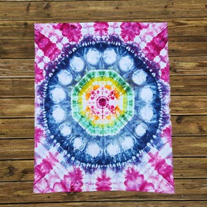 May include: A tie-dye textile with a circular mandala design. The center features a vibrant array of colors, including red, orange, yellow, and green. Surrounding the center are concentric circles of blue and white, with pink accents on the edges.