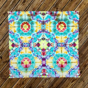 May include: A square, tie-dye textile with a vibrant, symmetrical pattern. The design features concentric circles in shades of turquoise, yellow, purple, and red. The fabric is laid on a wooden surface.
