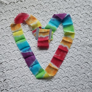May include: A rainbow tie-dye ribbon folded into a heart shape on a white crocheted surface.