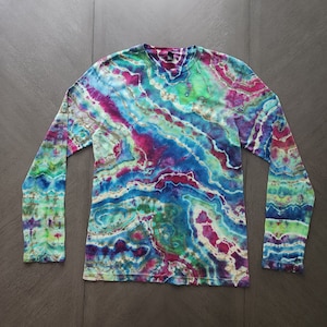 Pode incluir: Camiseta de manga comprida com um padrão tie-dye em tons de azul, verde, rosa e branco. A camiseta tem um decote redondo e um caimento solto.