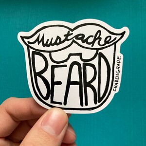 May include: A white sticker with a black outline of a mustache and beard. The text "Mustache Beard" is written in black inside the outline. The sticker is held in a hand.