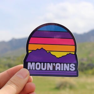 May include: A purple and black sticker with a sunset and mountain silhouette. The sticker says "Moun'tains" with the text "Grand Scale" below.