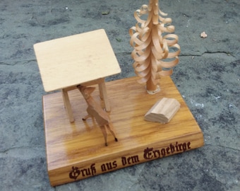 May include: A wooden miniature scene featuring a table, a deer, and a stylized tree. The base is inscribed with text. The scene is crafted from light-colored wood, with a glossy finish. The tree has a spiral design.