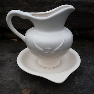 May include: White ceramic pitcher with a bird design and a matching saucer. The pitcher has a handle and a spout. The saucer has a slight curve to it.