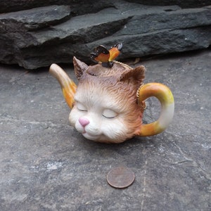 May include: A ceramic teapot shaped like a cat's head with a butterfly on top. The teapot is brown, white, and orange with a pink nose and closed eyes.