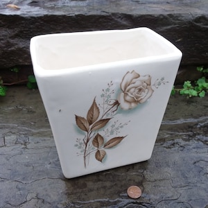 May include: White ceramic planter with a brown and green floral design featuring a single white rose.