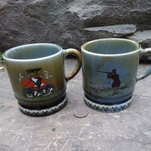 May include: Two green and white ceramic mugs with a Celtic design. One mug features a man carrying a sack on his back, the other mug features a man with a gun hunting.