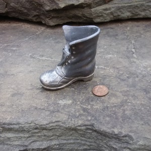 May include: A silver boot-shaped trinket with a lace-up design. The boot is sitting on a gray rock surface next to a penny.