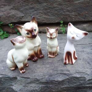 May include: Four ceramic cat figurines, two white with brown markings, one brown with white markings, and one white with brown markings and closed eyes. The figurines are sitting on a gray rock.