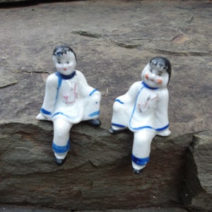 May include: Two porcelain figurines of children wearing traditional Chinese clothing. The figurines are painted in white, blue, and pink with black hair. They are sitting on a gray rock surface.