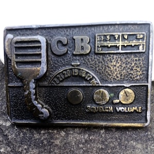 May include: A vintage-style belt buckle with a CB radio design. The rectangular buckle features a microphone, dials, and the letters "CB." The color is a mix of bronze and black, with the words "Squelch Volume" visible.