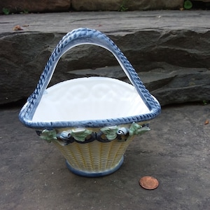 May include: A small, white ceramic basket with a blue handle and yellow woven design. The basket has a floral design with green leaves and blue berries.