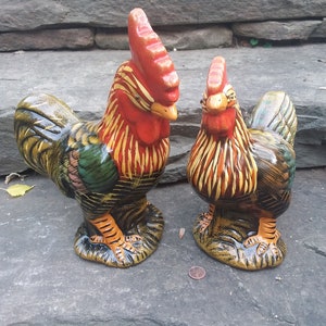 May include: Two ceramic rooster figurines, one larger than the other. The larger rooster is red, yellow, green, and black. The smaller rooster is yellow, orange, and brown. Both roosters have black eyes and orange feet.