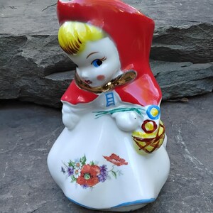 May include: A ceramic pitcher shaped like a girl wearing a red hooded cloak and holding a basket of flowers. The pitcher is decorated with floral details and has a white base.