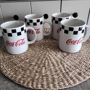 May include: Set of four white ceramic mugs with a black and white checkered pattern and the Coca-Cola logo in red. The mugs are on a woven straw placemat.