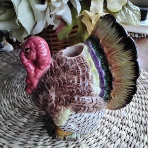 May include: A ceramic turkey figurine with a red head and a colorful tail. The turkey has a brown body with detailed feather patterns. The tail feathers display shades of yellow, purple, and black. This decorative piece is likely used for Thanksgiving or fall decor.
