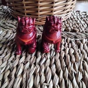 May include: Two red ceramic foo dog figurines with intricate details. The figurines are sitting on a woven straw mat.