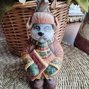 May include: A wooden figurine of a bear wearing a hat and scarf, holding a book. The bear is painted in shades of brown, green, and red.