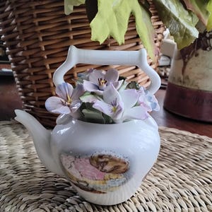 May include: White porcelain teapot with a floral design and a hedgehog illustration. The teapot is decorated with a bouquet of purple and white flowers.
