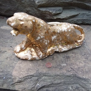 May include: A ceramic sculpture of a recumbent lion with a gold and white finish. The lion is lying down with its mouth open, and a penny is next to it for scale. The sculpture is on a gray stone surface.