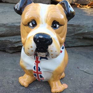 May include: A ceramic bulldog figurine featuring a tan and white coat with black ears and a black nose. The dog wears a white collar and a necktie decorated with a red, white, and blue Union Jack flag pattern. The surface has a glossy finish.