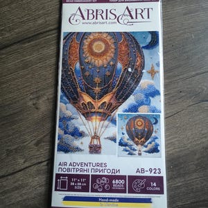 May include: Abris Art bead embroidery kit titled Air Adventures, model AB-923. The packaging features a hot air balloon design with a sun and stars, measuring 11 inches by 11 inches, containing 6800 beads in 14 colors on natural art canvas.