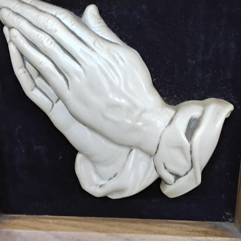 May include: A cream-colored sculpture of hands clasped in prayer. The hands are detailed with visible veins and wrinkles, set against a dark background. The sculpture is framed in a light-colored wood.