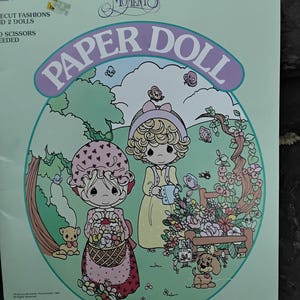 May include: A paper doll set featuring two Precious Moments dolls in a garden setting. The dolls are dressed in pink and yellow dresses with floral accents. The dolls are surrounded by flowers, butterflies, and a small dog. The paper doll set is titled "Precious Moments Paper Doll" and includes the text "Precious Moments, Incorporated 1982. All Rights Reserved. PRECIOUS MOMENTS is the trademark of Precious Moments, Incorporated."