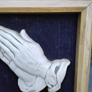 May include: A framed relief sculpture of clasped hands in a praying position. The hands are a light, off-white color, set against a dark blue background. The frame is made of light-colored wood.