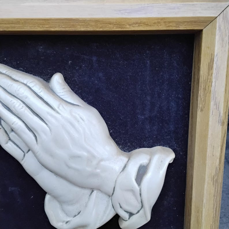 May include: A framed relief sculpture of clasped hands in a praying position. The hands are a light, off-white color, set against a dark blue background. The frame is made of light-colored wood.