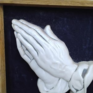 May include: A framed relief sculpture of two hands clasped in prayer. The hands are painted a light gray color and are set against a dark blue background. The frame is made of wood.