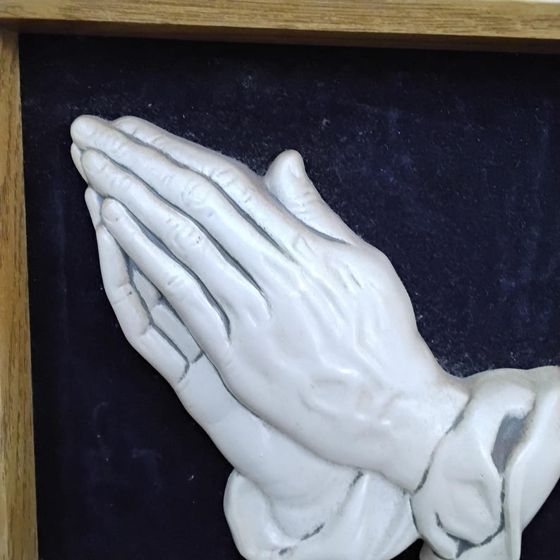 May include: A framed relief sculpture of two hands clasped in prayer. The hands are painted a light gray color and are set against a dark blue background. The frame is made of wood.