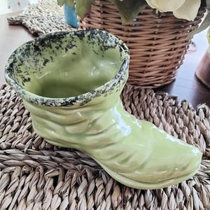 May include: A green ceramic boot planter with a black speckled glaze. The planter is sitting on a woven straw mat.
