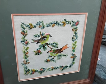 May include: A framed embroidery depicting two birds perched on a branch surrounded by a wreath of pine branches and pine cones. The birds are green, orange, and brown. The background is a light green.