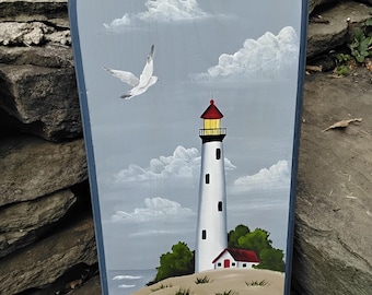 May include: A hand-painted wooden sign depicting a lighthouse, a small house, and two seagulls flying in a blue sky with white clouds. The sign is painted in a whimsical style with a light blue background and white accents.