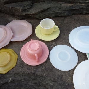 May include: A set of pastel-colored teacups and saucers. The set includes a pink cup and saucer, a yellow cup and saucer, a white saucer, a light blue saucer, a yellow saucer, and a pink saucer. The saucers are all different shapes and sizes.
