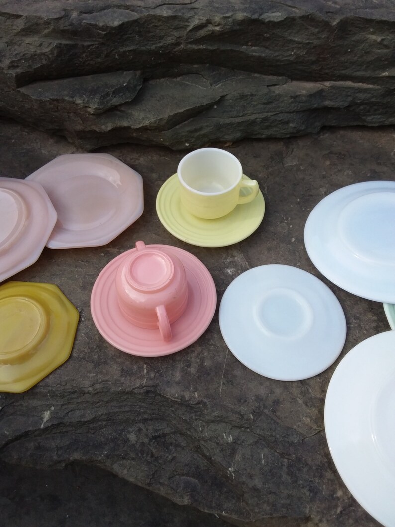 May include: A set of pastel-colored teacups and saucers. The set includes a pink cup and saucer, a yellow cup and saucer, a white saucer, a light blue saucer, a yellow saucer, and a pink saucer. The saucers are all different shapes and sizes.