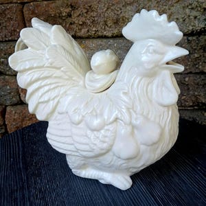 May include: A cream-colored ceramic rooster-shaped pitcher with a glossy finish. The pitcher features detailed molded feathers, a comb on top, and a decorative cluster of pears embossed on the side. It has an open beak spout and a lid.