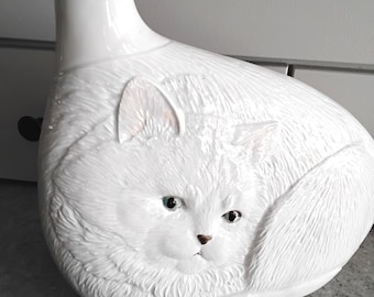 Puede incluir: Un jarrón de cerámica blanca con forma de gato enroscado, con detalles de pelaje texturizado, ojos azules y una pequeña huella de pata rosa en la base. El jarrón tiene una abertura en la parte superior con un ramo de flores rosas y blancas.