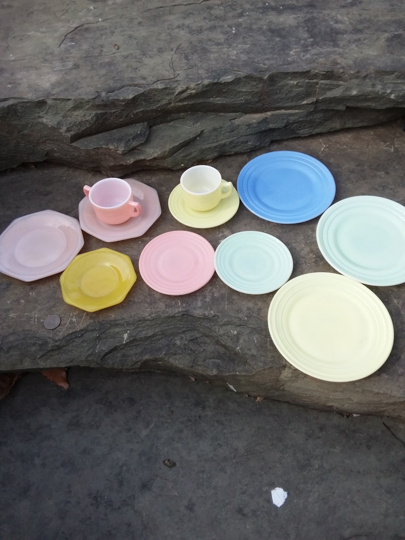 May include: A set of pastel-colored teacups and saucers, including a light blue saucer, a light green saucer, a yellow saucer, a pink saucer, a light yellow saucer, and a pink saucer. The teacups are white with a pink rim.