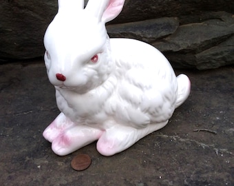 May include: A white ceramic rabbit figurine with pink accents on the inner ears, nose, and paws. The rabbit is in a seated position with textured fur detailing. A copper-colored penny is placed on the ground next to the rabbit for scale.