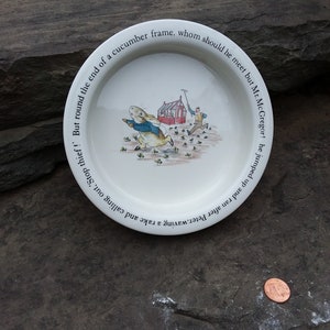 May include: White ceramic plate with a black border and a Peter Rabbit illustration. The plate has a quote around the edge that reads: "and ran after Peter. waving a rake and calling out, Stop thief! But round the end of a cucumber frame. whom should he meet but Mr. McGregor! He jumped up and"