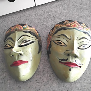 May include: Two hand-painted wooden masks featuring pale green faces, arched black eyebrows, almond-shaped eyes, and red lips. The masks are adorned with intricate floral-patterned headpieces in shades of orange, yellow, and black.