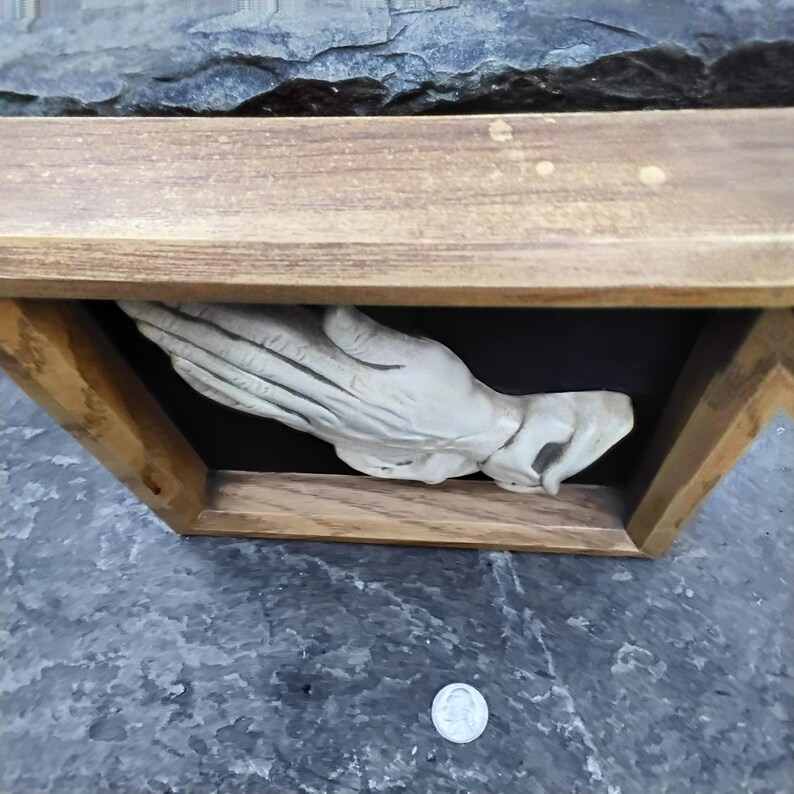 May include: A wooden framed relief sculpture of a hand, painted in a light color. The hand is cupped, as if holding something. The frame is made of light brown wood. The background is black.