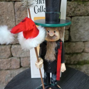 May include: A wooden nutcracker figure wearing a black top hat with a green brim and a black coat. The figure has a brown beard, holds a wooden staff topped with a red and white fabric hat, and stands in front of a box labeled Hei Colle.