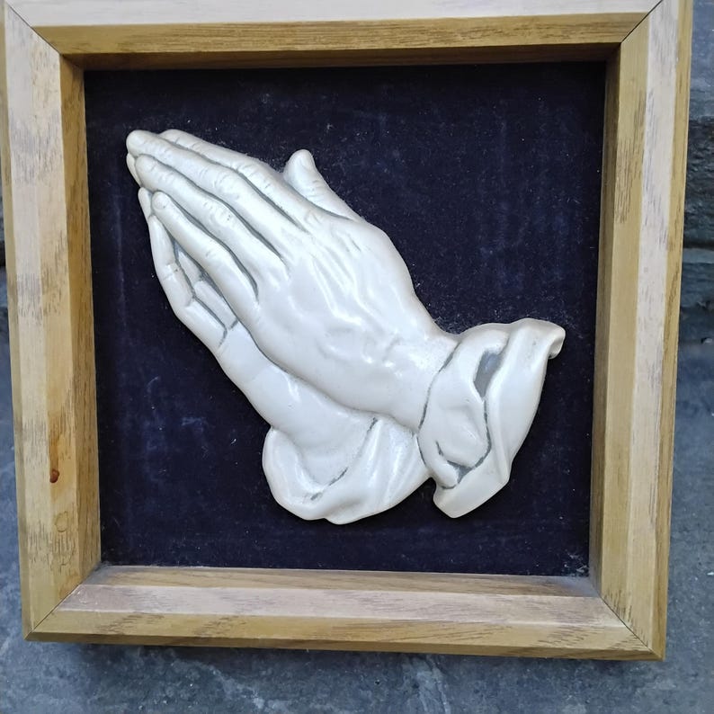 May include: A framed relief sculpture of praying hands. The hands are white and detailed, set against a dark blue background. The frame is made of light-colored wood, adding a natural touch to the artwork. The sculpture is a symbol of faith and devotion.