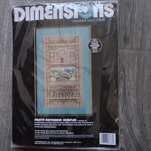 May include: A Dimensions counted cross stitch kit in a black package. The kit features a sampler design with a floral and house motif, and the text "ABCDEFGHIJ KLMNOPORS TUVWXYZ". The kit includes a colour-accented chart and is fun to stitch.