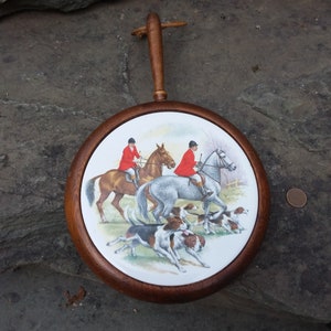May include: A round wooden wall plaque with a ceramic insert featuring a hunting scene. Two riders in red jackets are on horseback with hounds running in front of them. The plaque has a wooden handle.