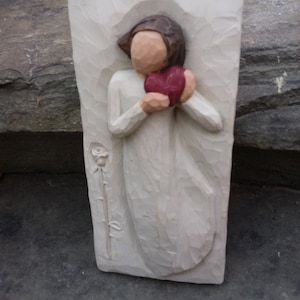 May include: A white wooden figurine of a person holding a red heart in their hands. The figurine is carved in a simple style and has a rustic look. The person is wearing a long white robe and has a simple face. The figurine is standing on a white base.